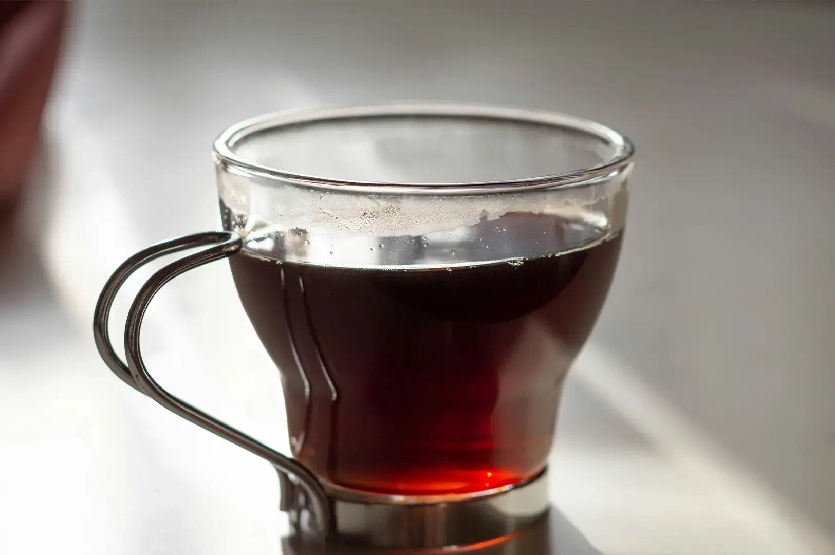 A close-up photograph displays a modern, clear glass teacup filled with dark, amber-colored black tea. The teacup features a distinct, sleek metallic handle and base that accents its curved glass design. Sunlight catches the rim of the cup, casting a warm glow through the translucent liquid, while tiny condensation droplets line the glass just above the tea surface against a softly blurred, neutral background.