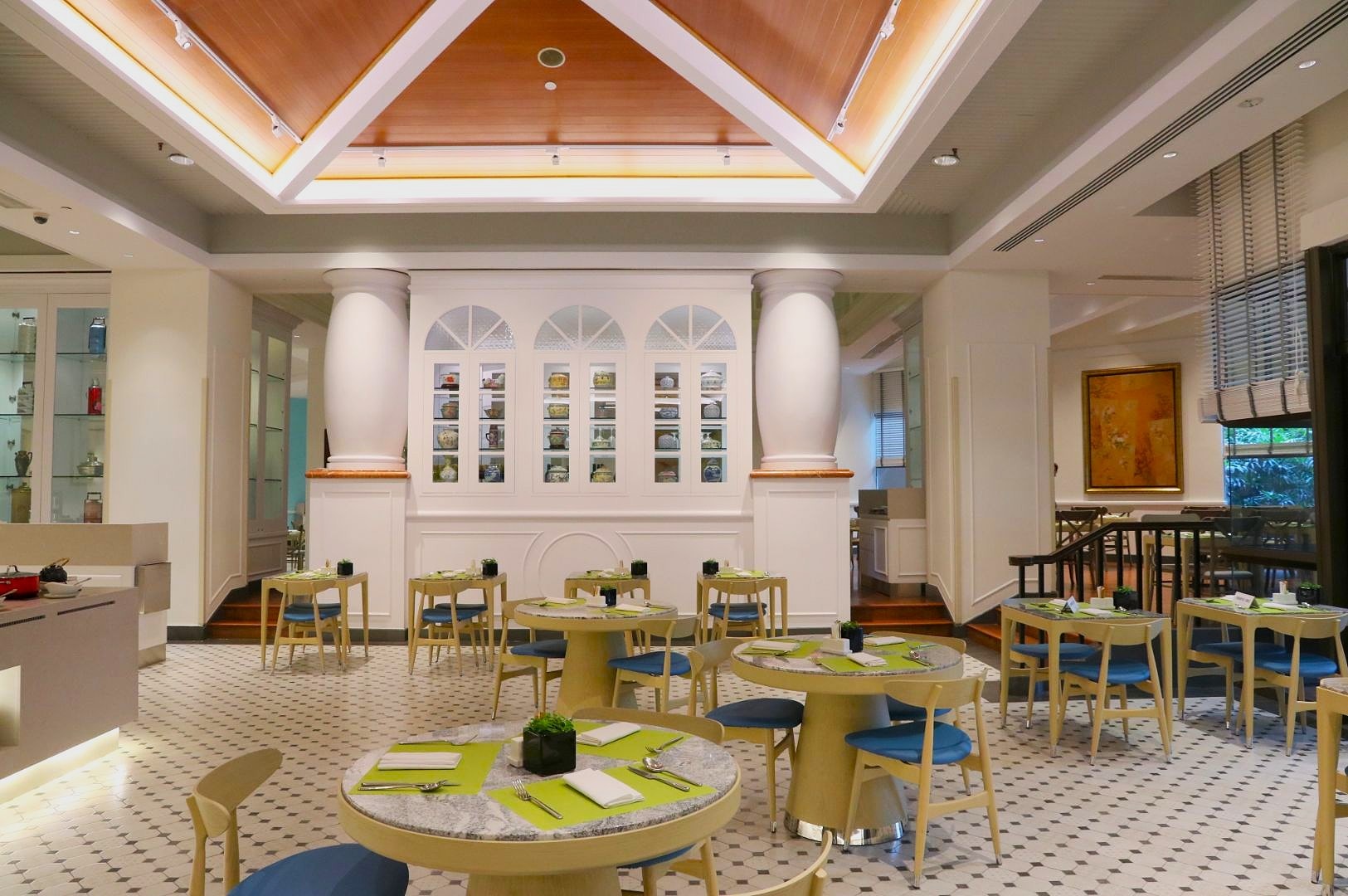 Chic restaurant interior with round tables, green placemats, and blue chairs. Soft lighting enhances the elegant, cozy ambiance. White pillars and wall art add style.