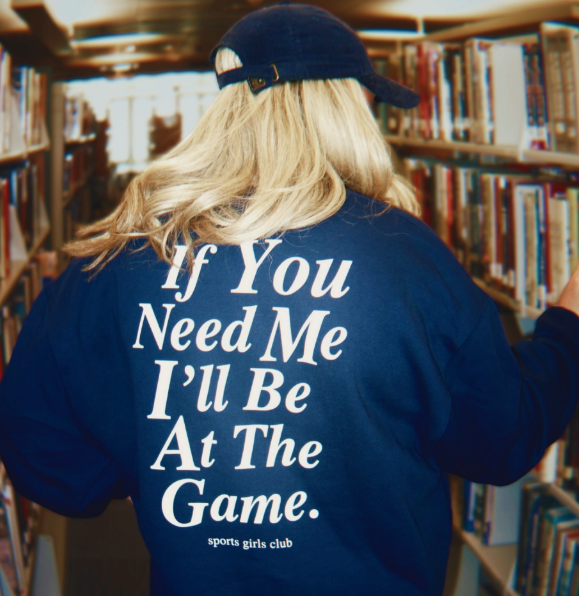 blond woman wearing navy crewneck sweatshirt that says if you need me I'll be at the game" in a library