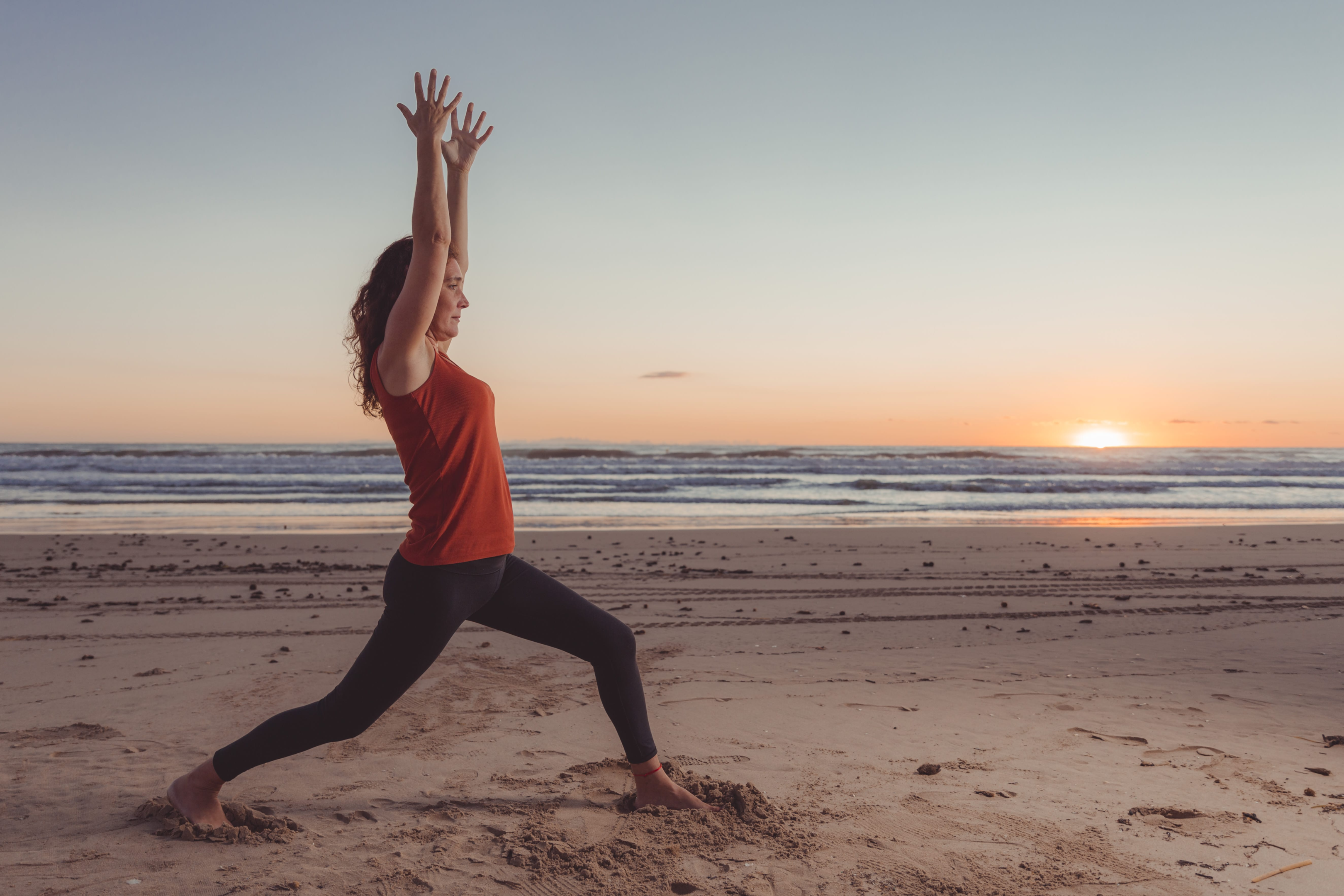 woman doing yoga with good posture.  She is standing on a beach at sunrise wearing black pants and a red top.