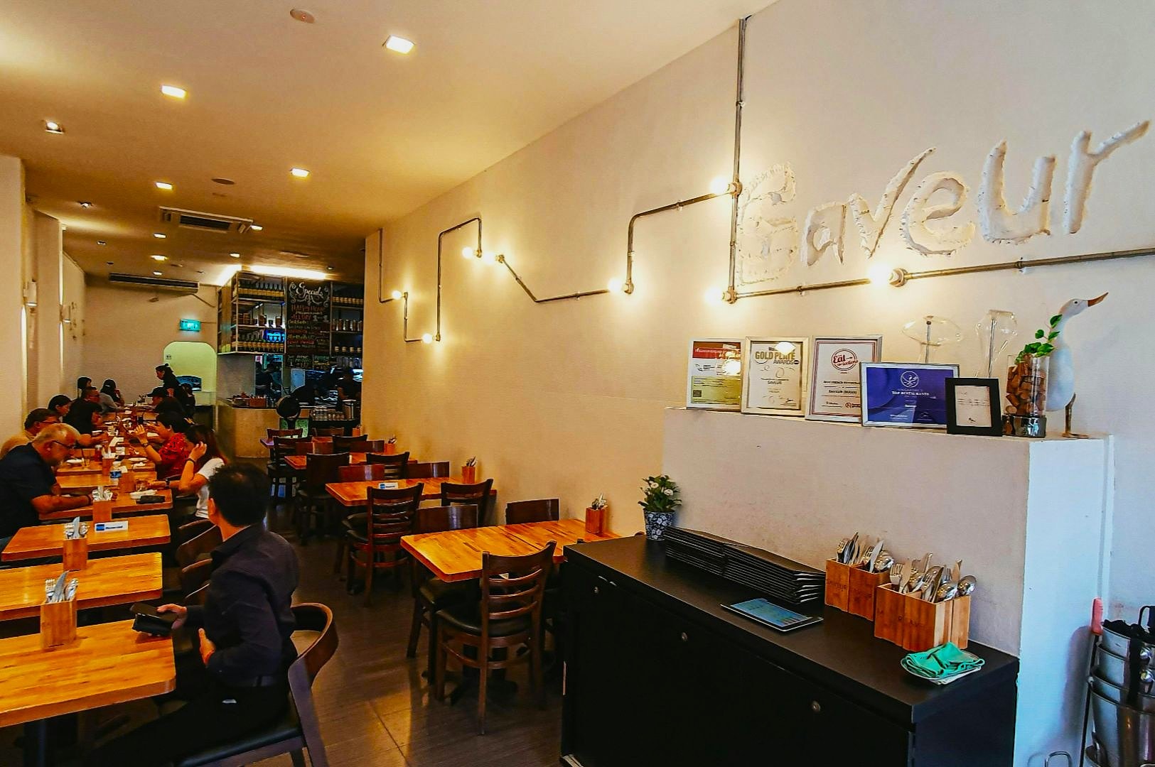 Casual dining restaurant interior with wooden tables and chairs. People are seated, talking and eating. Awards and lights decorate the white wall. Cozy ambiance.