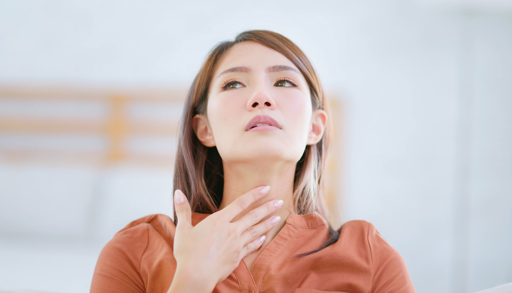 Kvinne holder seg på halsen og ser opp med bekymret uttrykk, muligens på grunn av sår hals eller ubehag.