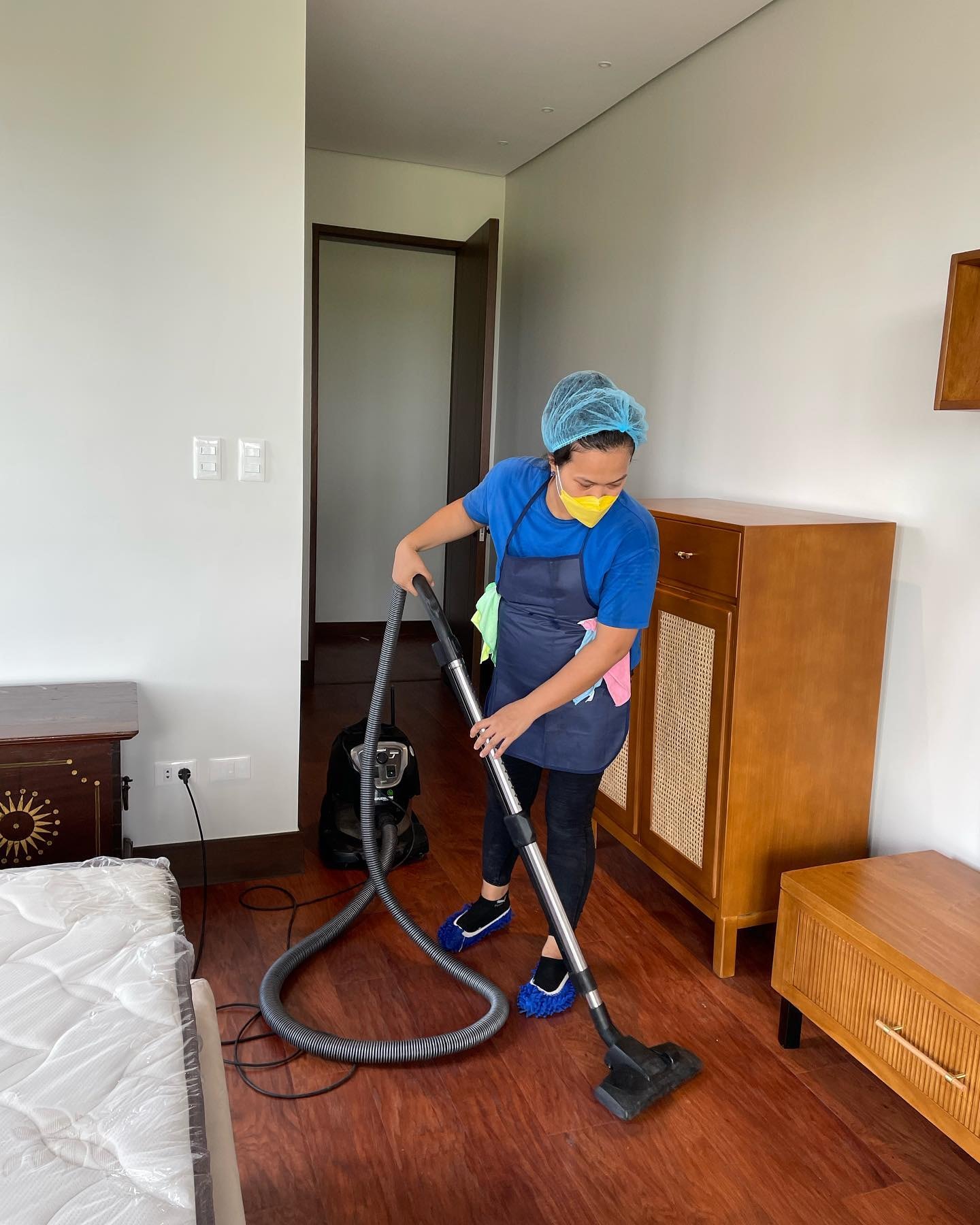 Person wearing a hair net, face mask, and apron vacuuming a wooden bedroom floor as part of an end of tenancy deep clean — with cleaning cloths tucked into apron pockets and a vacuum cleaner in use.