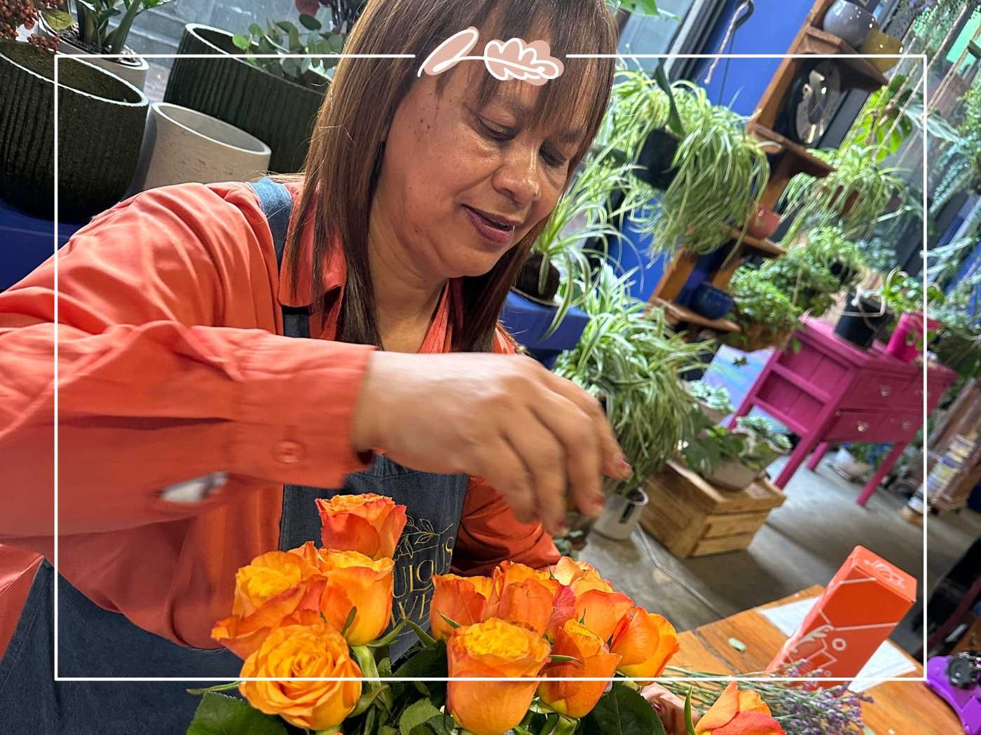 A woman in an orange shirt arranging bright orange roses in a vase, surrounded by a cozy shop setting filled with plants and colorful furniture. Fabulous Flowers and Gifts.