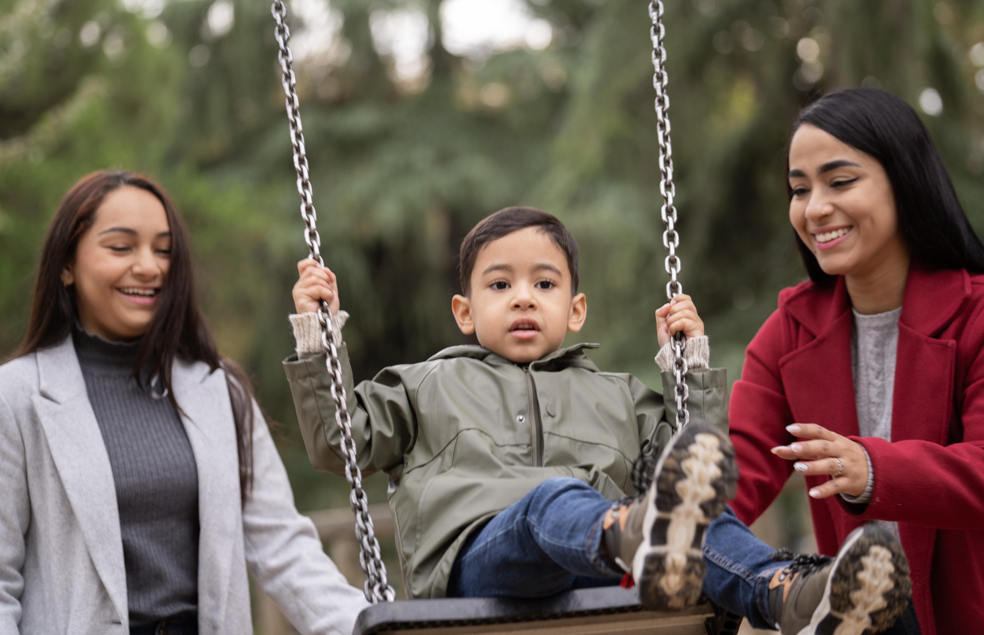 A young child in an olive green jacket sits on a swing, holding the silver chains.
