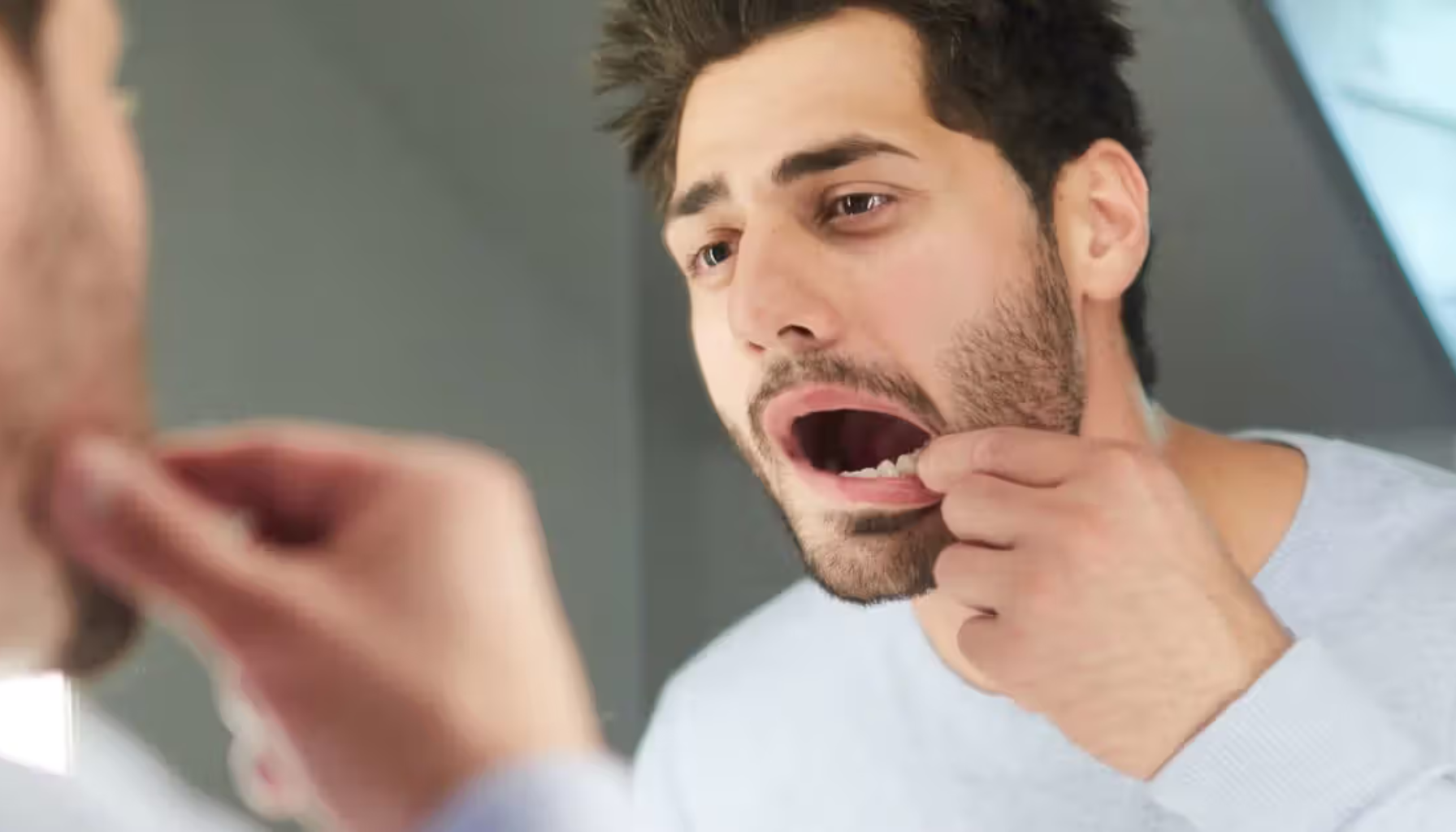 Mann ser i speilet med åpen munn, undersøker munnen for mulig problem eller ubehag.