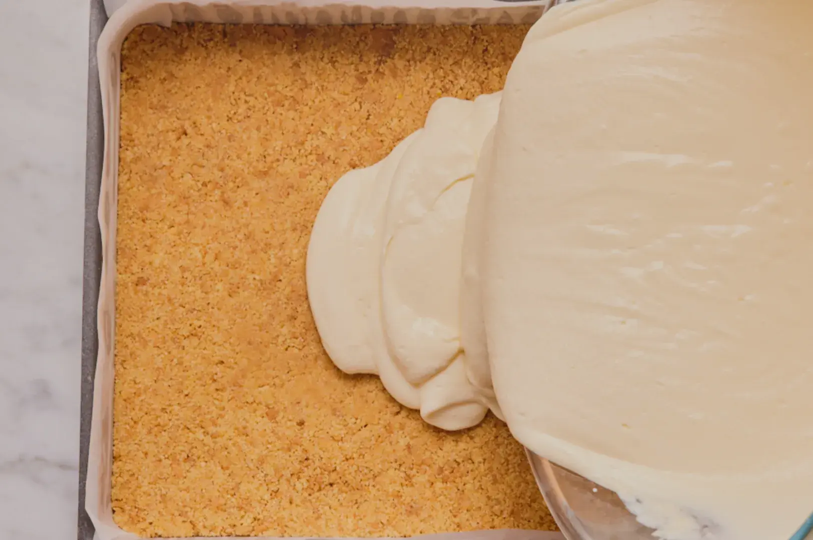 A parchment-lined rectangular pan holds a golden crumb crust as thick white cream is poured from a bowl, ready to be spread for a homemade dessert.