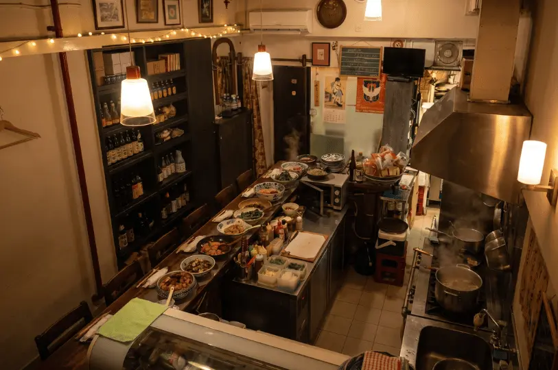 Overhead view of a cozy, narrow Japanese restaurant interior featuring a long wooden bar counter filled with prepped dishes, a busy cooking line, and shelves stocked with sake bottles.