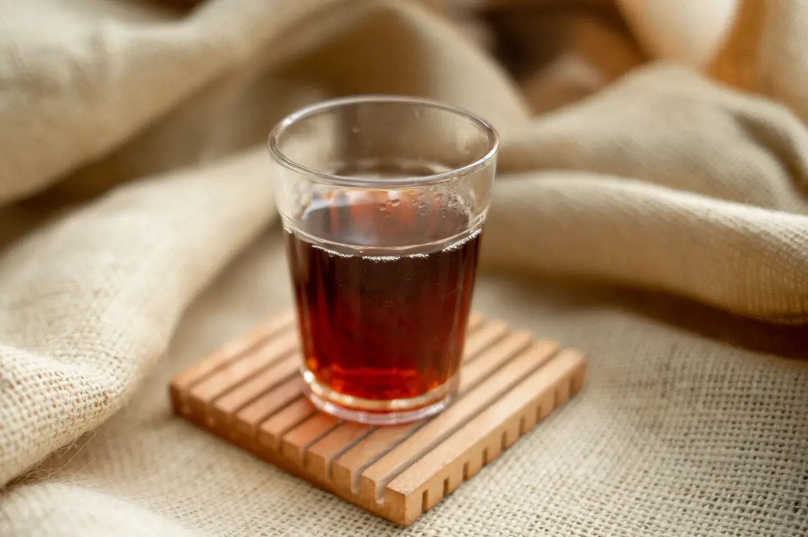 Clear glass filled with dark tea resting on a square slatted wooden coaster against folded neutral beige fabric.