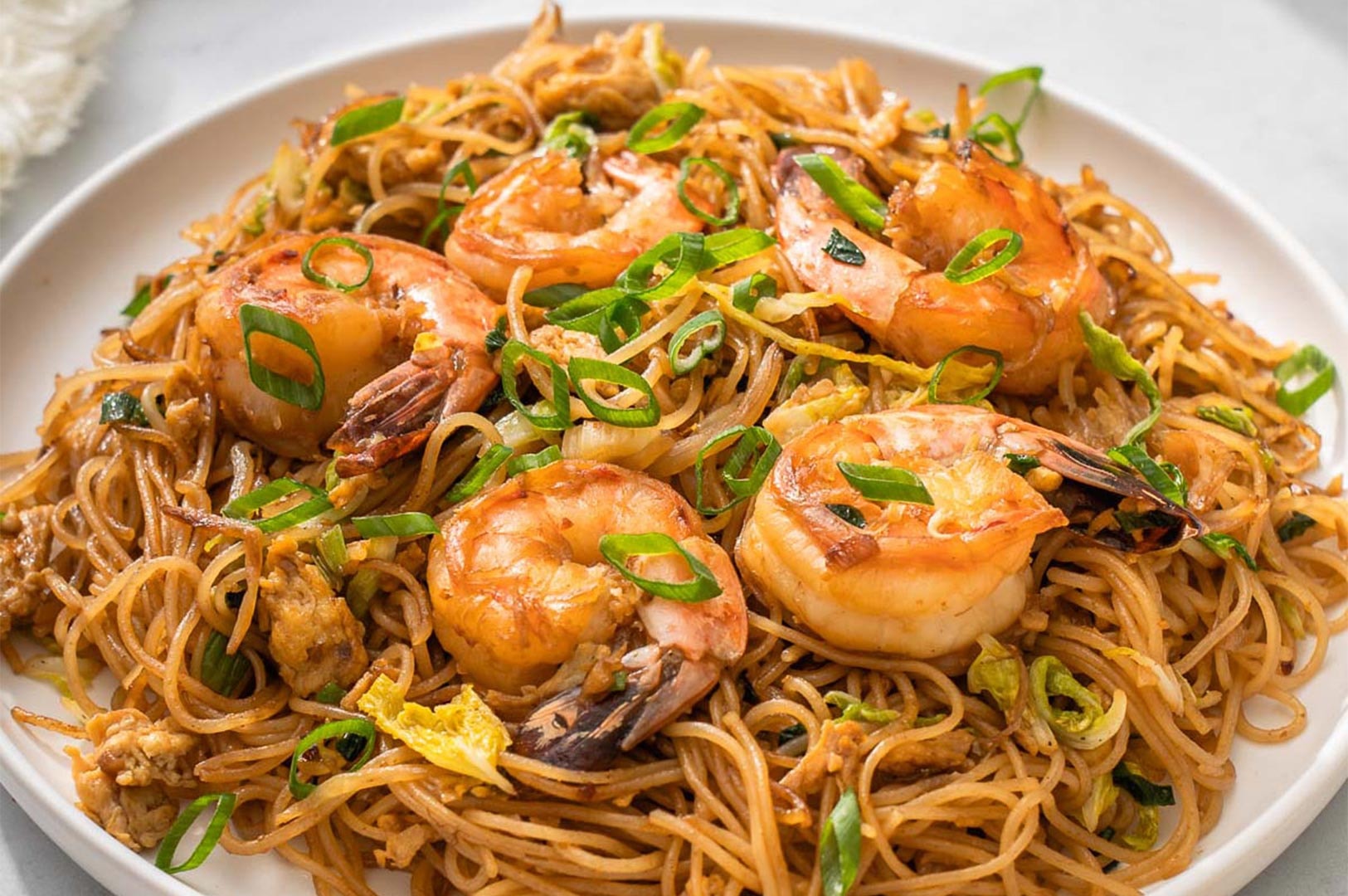 A close-up of savory fried rice noodles seasoned with soy sauce, topped with large grilled prawns and scallions.