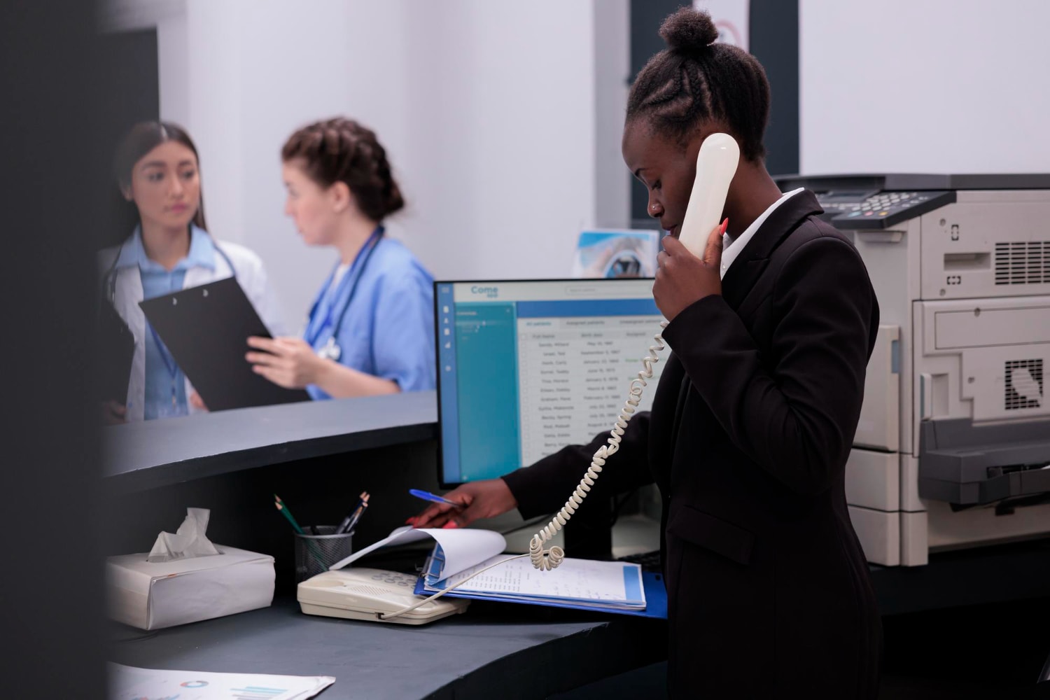 A receptionist in a healthcare setting using a corded phone while checking documents, with two medical professionals in the background. This highlights Hosted PBX Solutions optimizing communication for healthcare professionals.