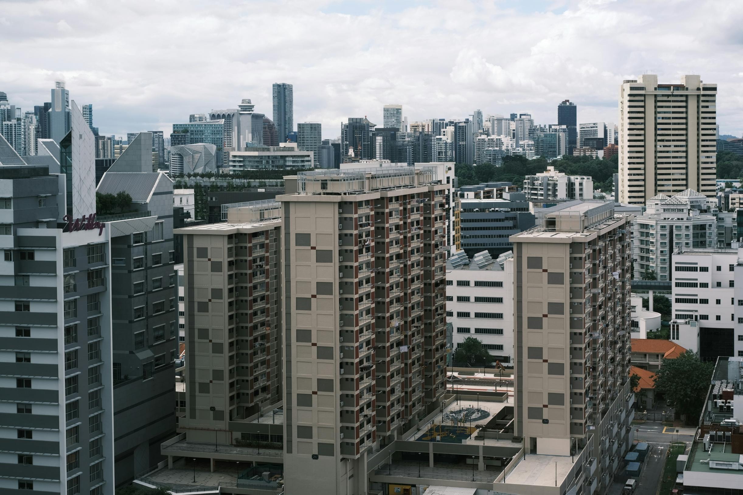 A cluster of high-rise apartment buildings with beige facades dominates the foreground, standing alongside a modern grey structure with an angular roofline. The backdrop features a dense and expansive city skyline, where numerous skyscrapers and commercial buildings stretch towards the horizon under a cloudy sky.