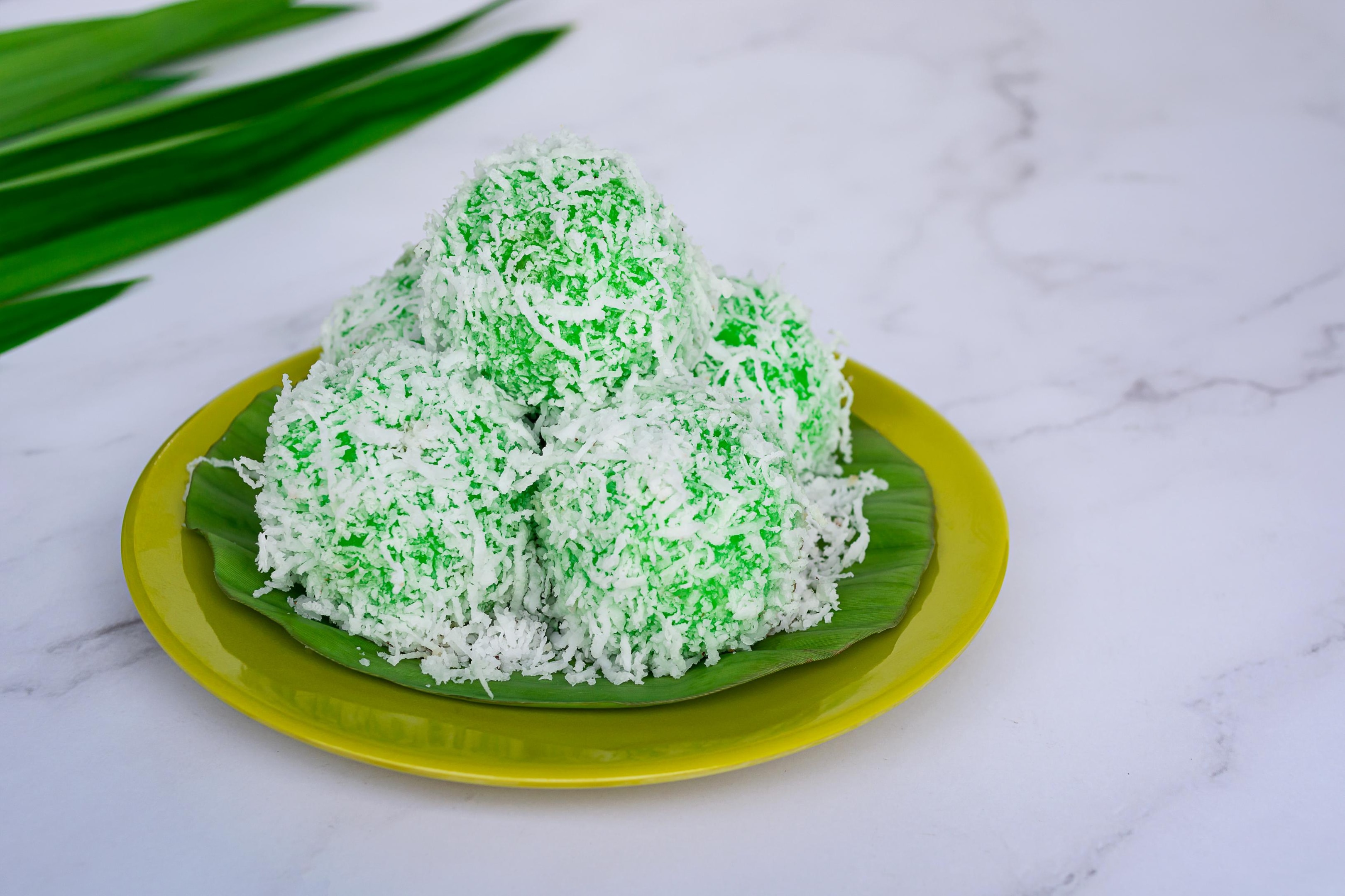 This photograph features a stack of five round, bright green Malaysian/Indonesian dessert balls, known as Klepon or Onde-onde, heavily coated in shredded white coconut.