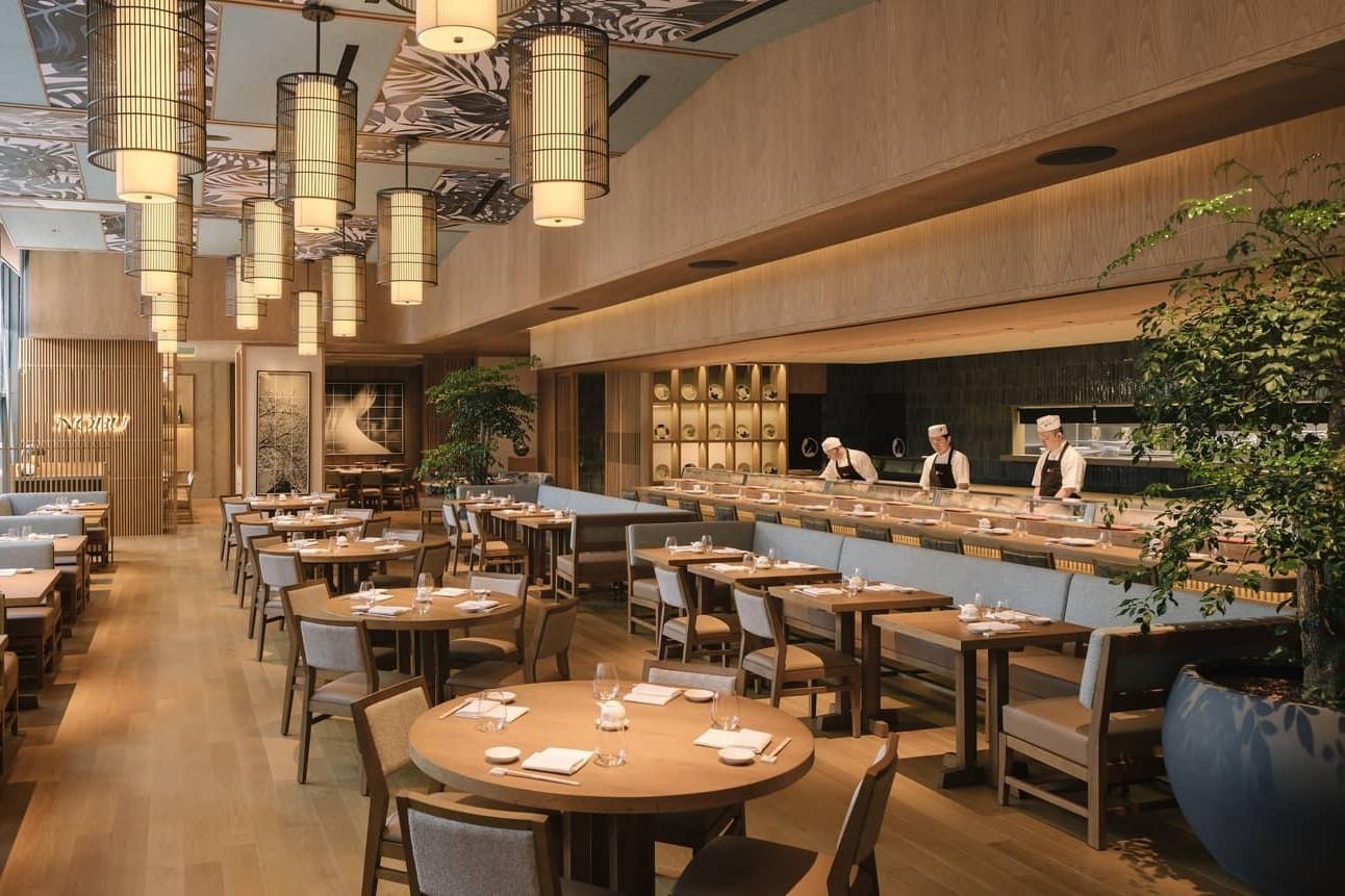  This airy, high-end restaurant features a spacious dining area with round wooden tables and a long sushi counter where chefs are at work. The interior is accented by light wood paneling, decorative botanical ceiling tiles, and a series of tall, cylindrical birdcage-style lanterns that provide soft, warm illumination.
