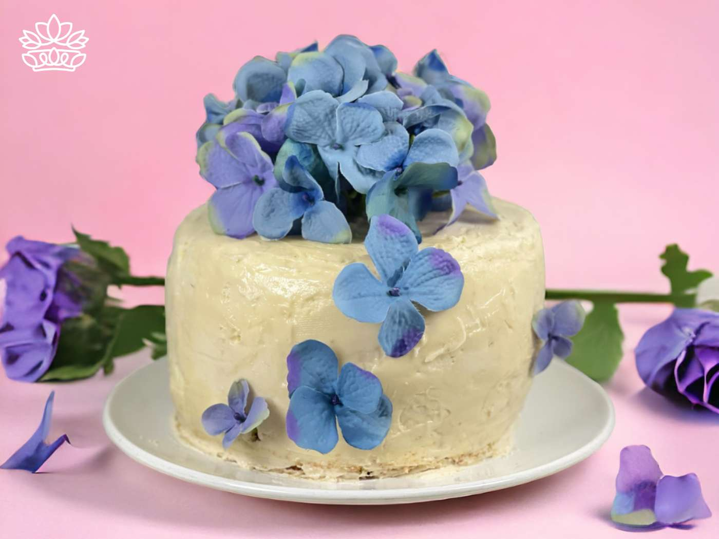A delicate cake adorned with a cascade of blue hydrangea blossoms, set against a soft pink backdrop, a testament to the artistic bakery paired with floristry at Fabulous Flowers and Gifts.