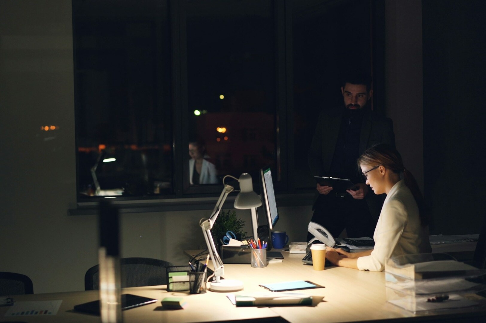 A woman in an office works at a desk under a desk lamp, with papers and a computer. A man stands behind her with a tablet. The scene is dimly lit, focused, and serious.