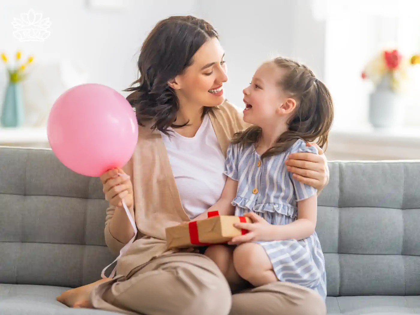 Mother and daughter sitting on a couch, holding a pink balloon and a gift box. Fabulous Flowers and Gifts. Collection: Women's Day.