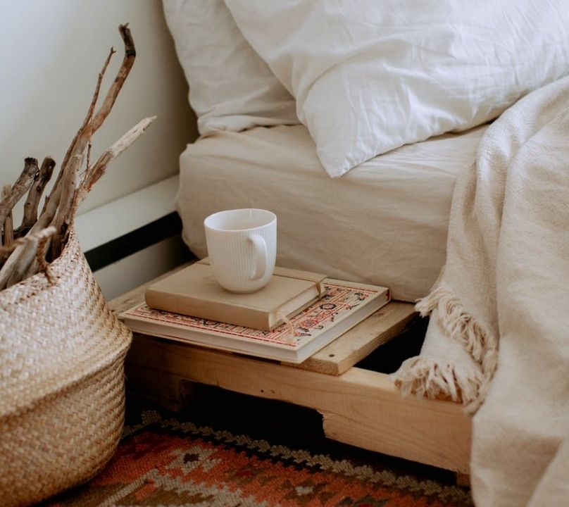 Comfortable cozy bedroom with bed wooden shelves with book and cup while wicker basket with sticks placed on ethnic styled rug
