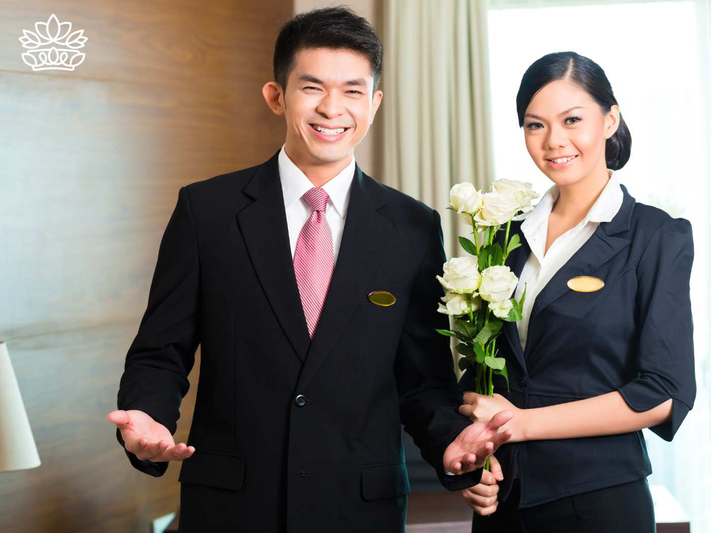 Welcoming hotel staff, a male and a female, in professional attire, greet guests warmly. The man, wearing a suit and red tie, gestures invitingly, while the woman holds a bouquet of fresh white roses, symbolizing exceptional hospitality and care. Fabulous Flowers and Gifts for Guest House and Hotel Flowers"
