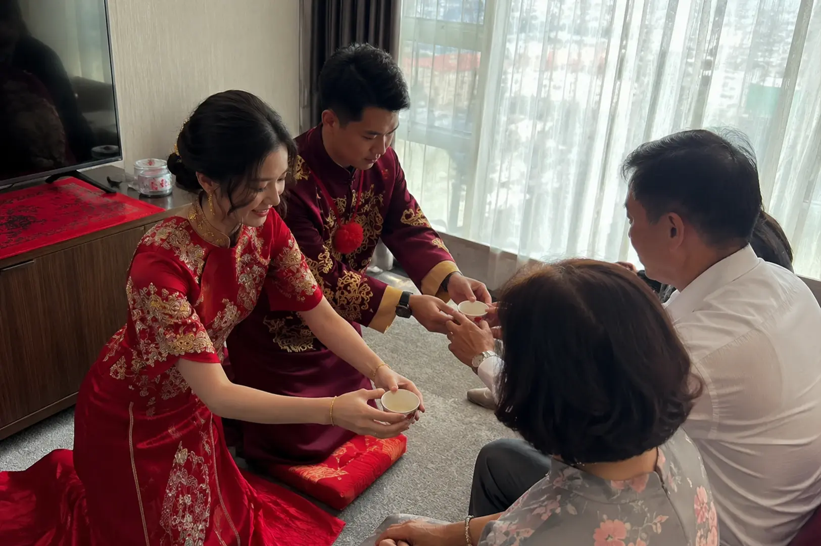 A bride and groom are kneeling on red cushions on a gray carpeted floor, bowing slightly as they offer tea cups to an older couple seated in front of them. The bride is on the left, wearing an elegant bright red Qun Kwa adorned with intricate gold and silver embroidery, her hair styled in an updo with gold hairpins. The groom knees beside her in a deep burgundy satin robe with gold embroidery and a red fabric rosette across his chest. They hand small white teacups to a seated man in a white dress shirt and a woman with short dark hair wearing a gray floral blouse, set against a backdrop of large windows with sheer white curtains.