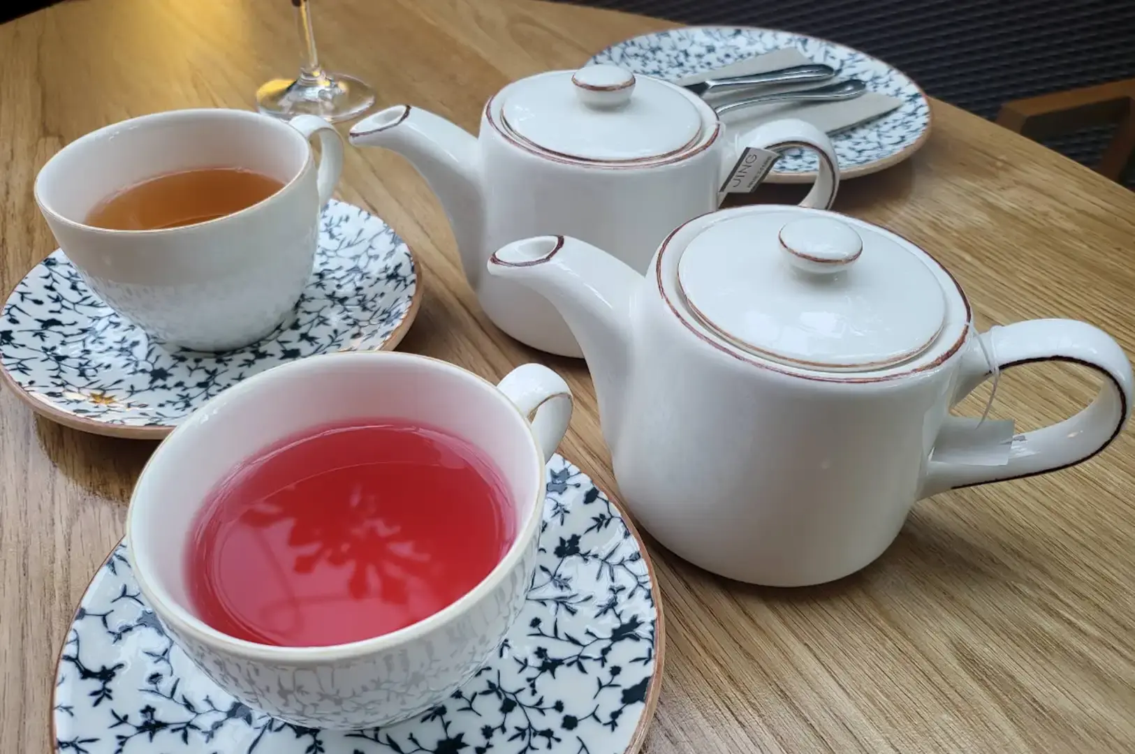 A table setting featuring two white ceramic teapots with brown-rimmed lids and spouts, arranged with two teacups on blue-and-white floral saucers—one containing light golden tea and the other a bright pink herbal infusion.