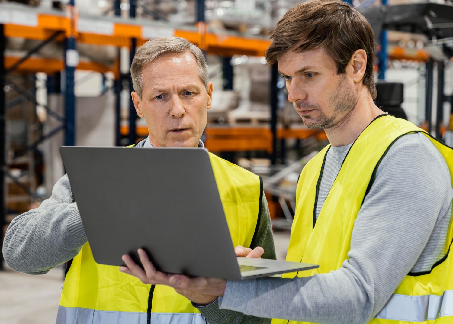 Two men in high-visibility vests stand in a warehouse aisle, closely reviewing data on a laptop. Through UCaaS, they leverage cloud telephony and unified communications for enhanced logistics, inventory tracking, and instant collaboration via cloud-based communication platforms.