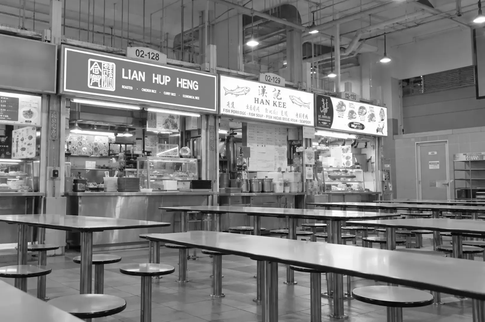 A traditional hawker centre seating area with rows of tables and stools facing several local food stalls.