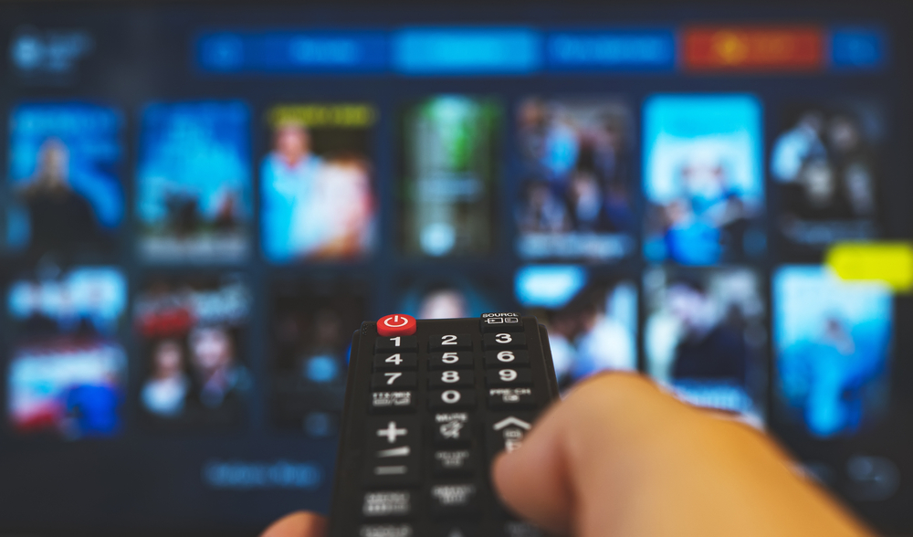 a hand holding a TV remote points it towards a streaming service interface in the background