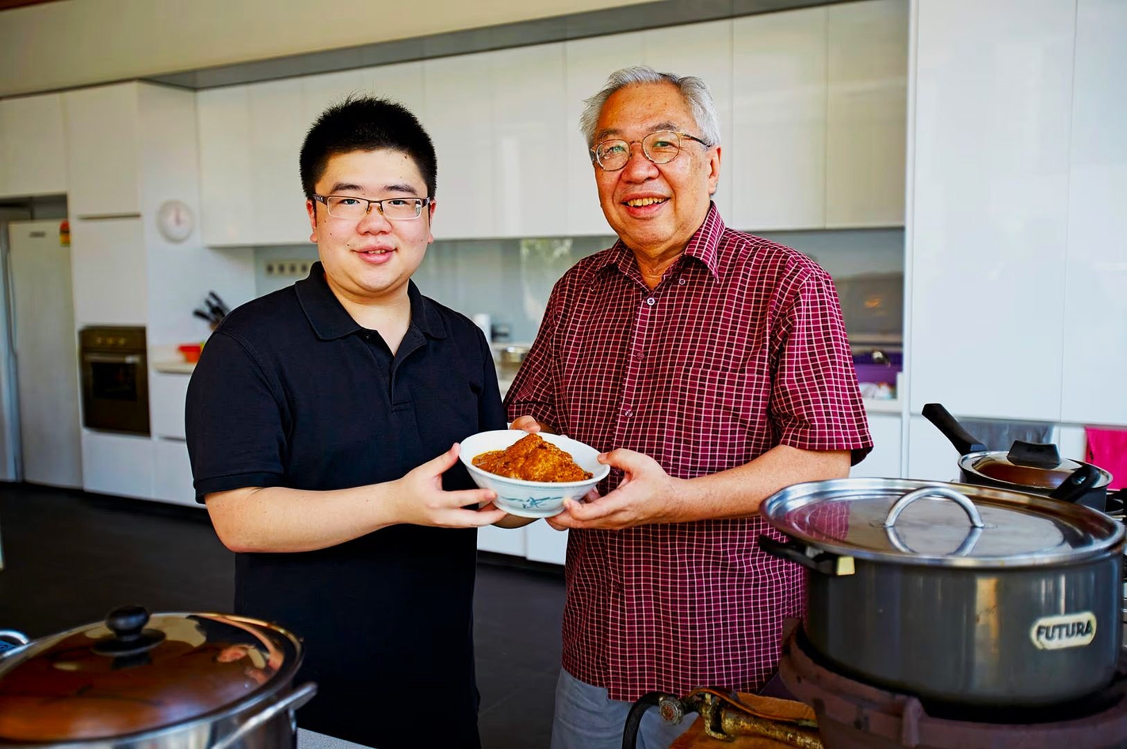 Two smiling men stand in a modern kitchen, holding a bowl of food together. Cookware is visible on the countertop, creating a warm, homey atmosphere.