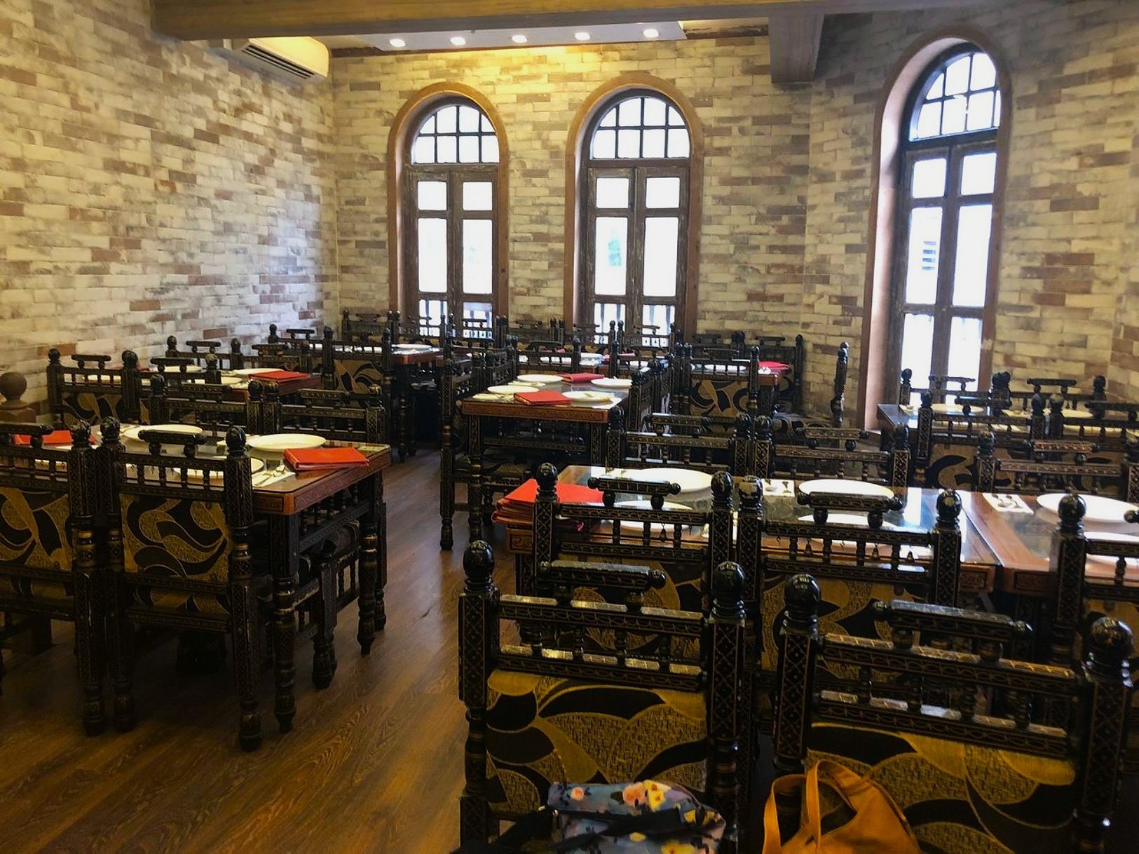 Cozy restaurant interior with ornate dark wooden chairs and tables set with red menus. Large arched windows let in natural light, enhancing the warm brick walls.