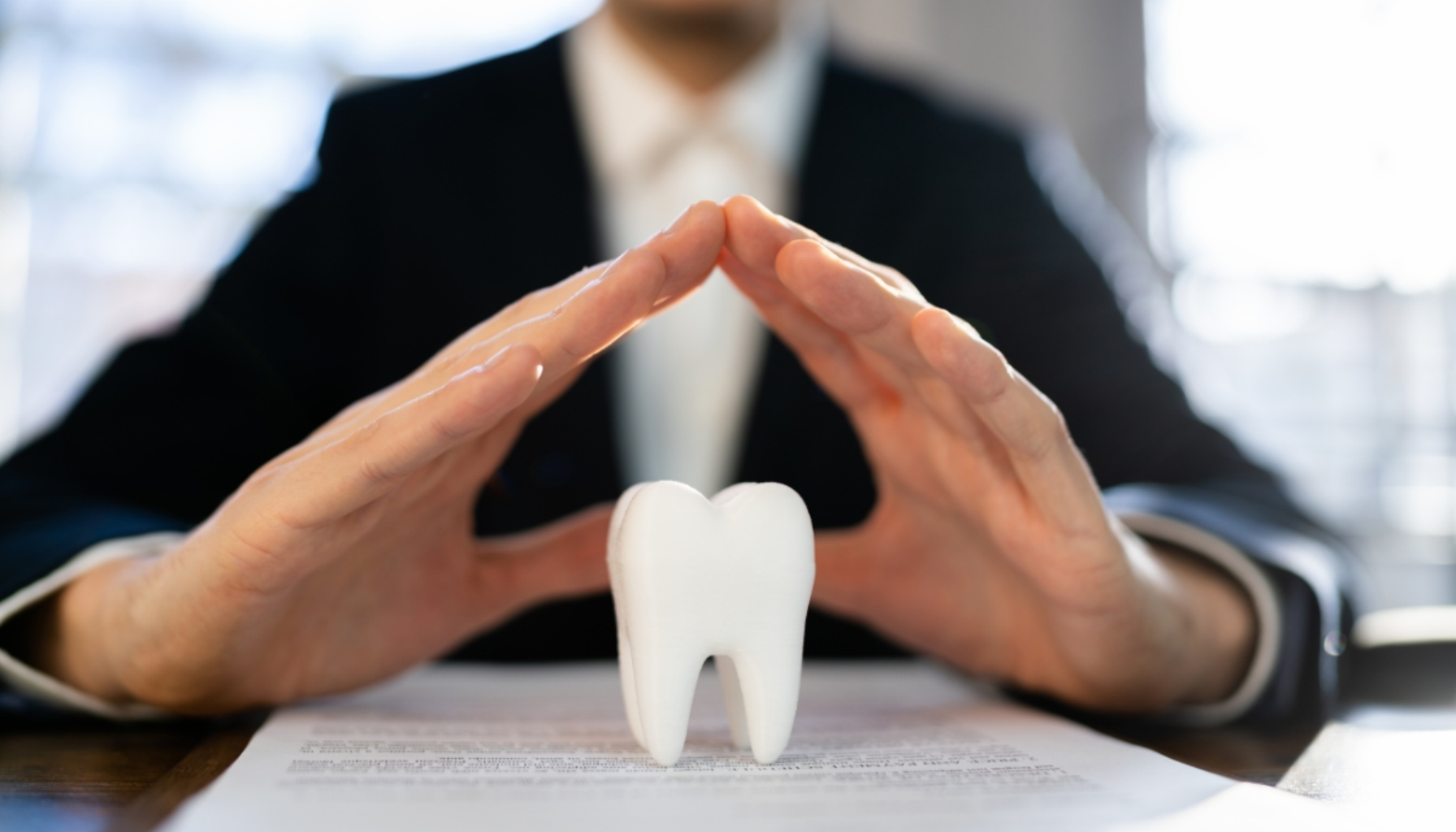 Tannhelseforsikring: En trygghet for økonomien 11 Person i dress former et beskyttende tak med hendene over en tannmodell på bordet.