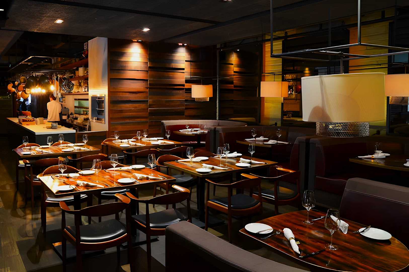 Modern restaurant interior with wooden tables, neatly set with white plates and wine glasses. Warm lighting creates a cozy, elegant atmosphere.