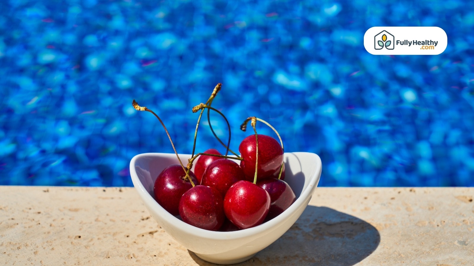 Fresh cherries in white bowl beside a bright blue swimming pool.
