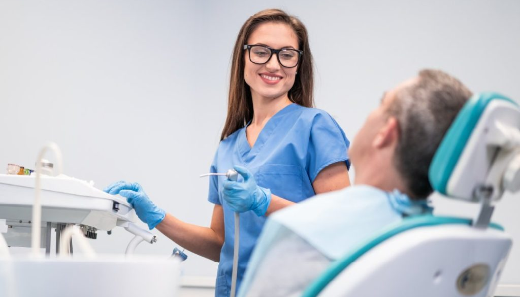 Tannpleier i blå uniform smiler og snakker med pasient som sitter i tannlegestol under rutinemessig kontroll.