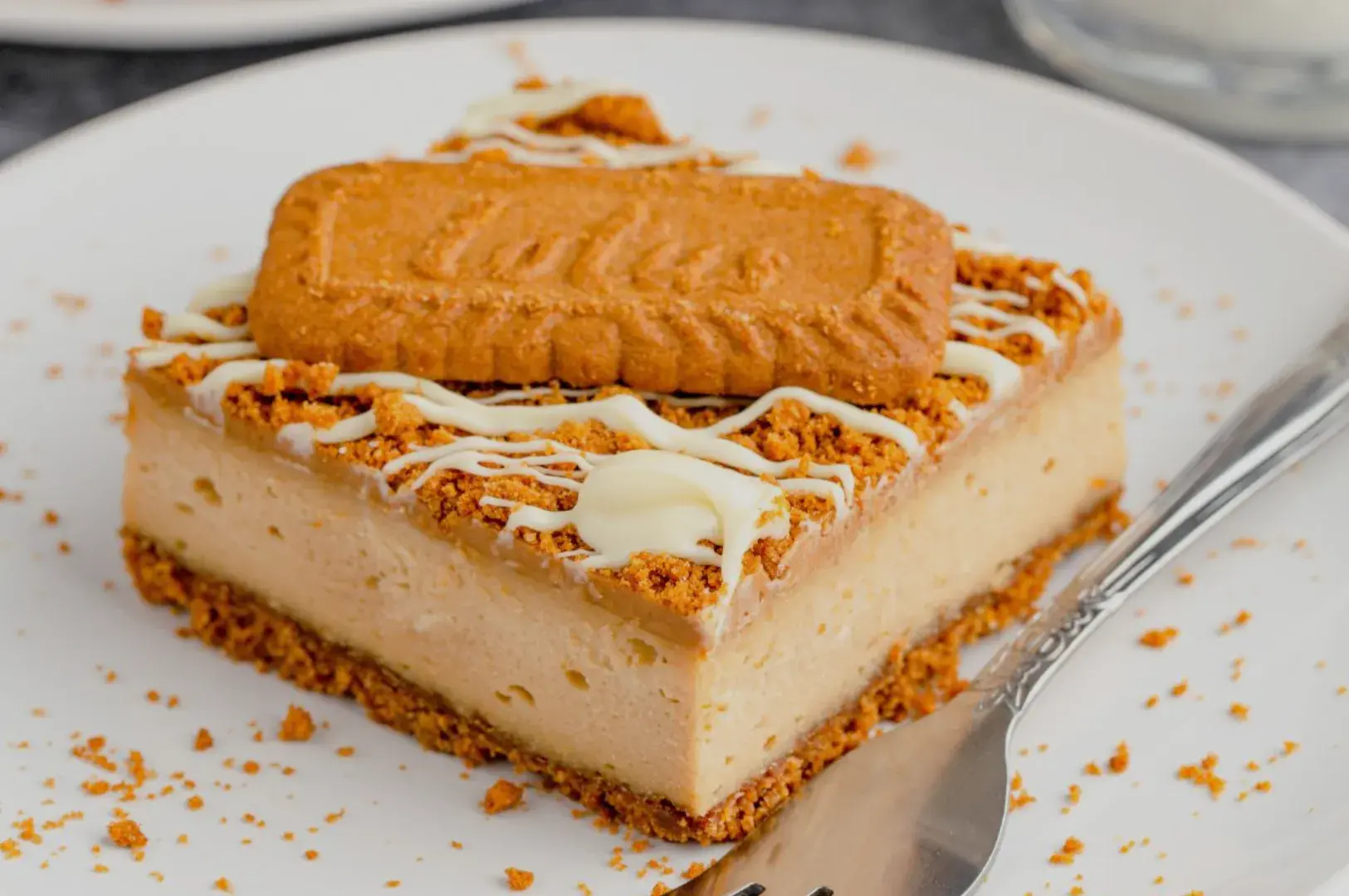 A square slice of dessert topped with a cookie and drizzled with white chocolate, sitting on a white plate with some crumbs around it.