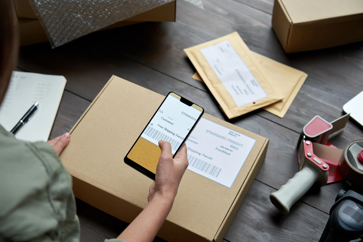 A hand holds a smartphone scanning a shipping label on a cardboard box, surrounded by materials like bubble wrap, envelopes, and tape dispenser.