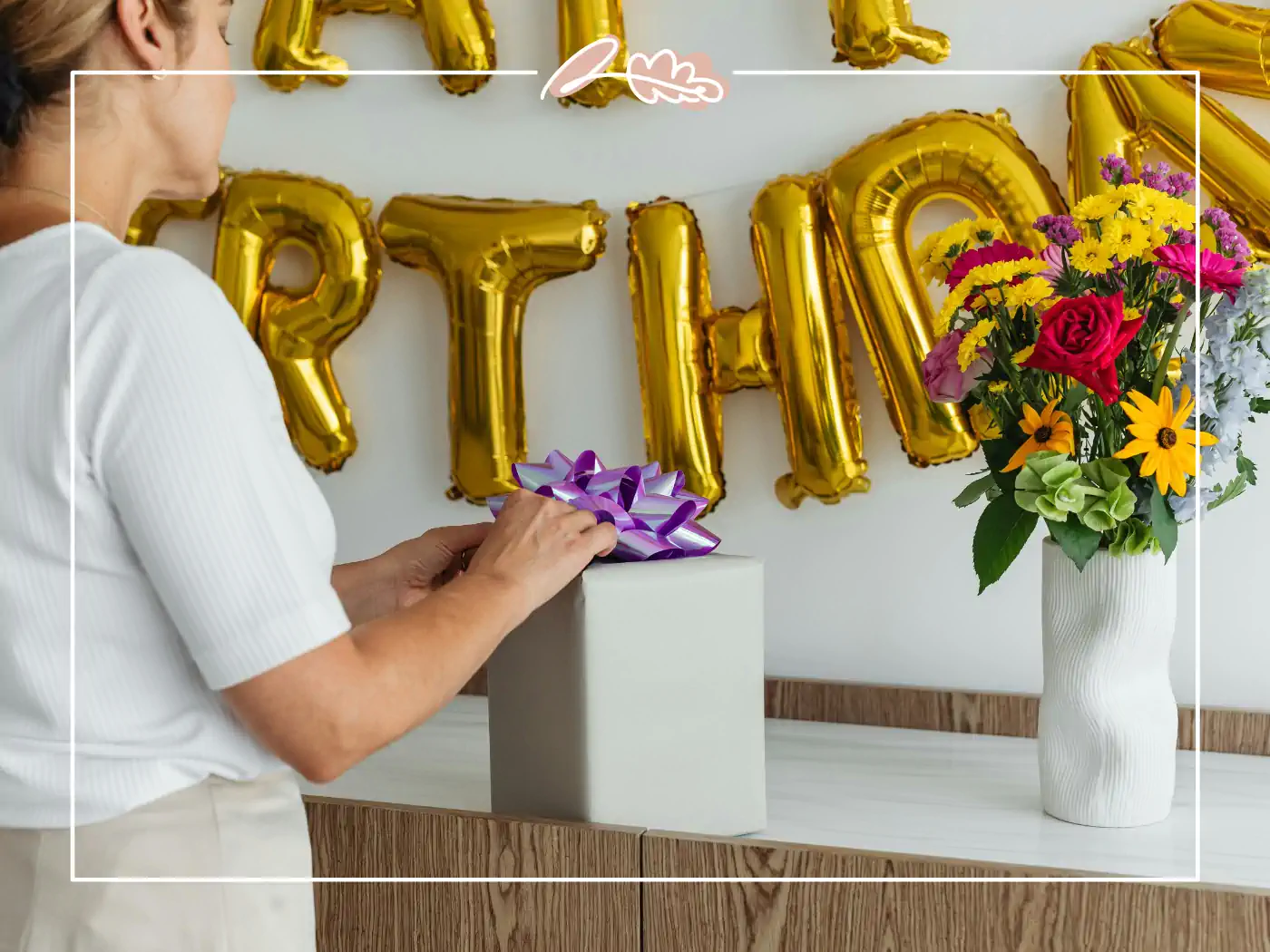 A woman in white placing a gift box with a purple ribbon on a table, with birthday decorations in the background. Fabulous Flowers and Gifts.