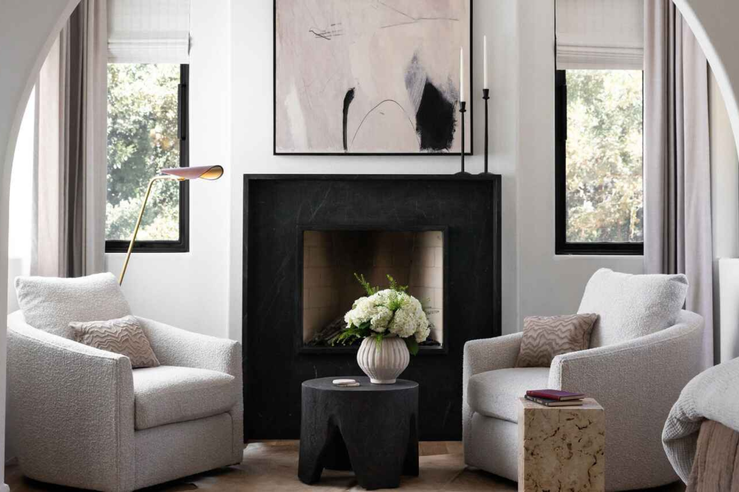 Intimate seating area featuring two boucle swivel chairs, a black fireplace, abstract artwork, and soft arched architectural framing.