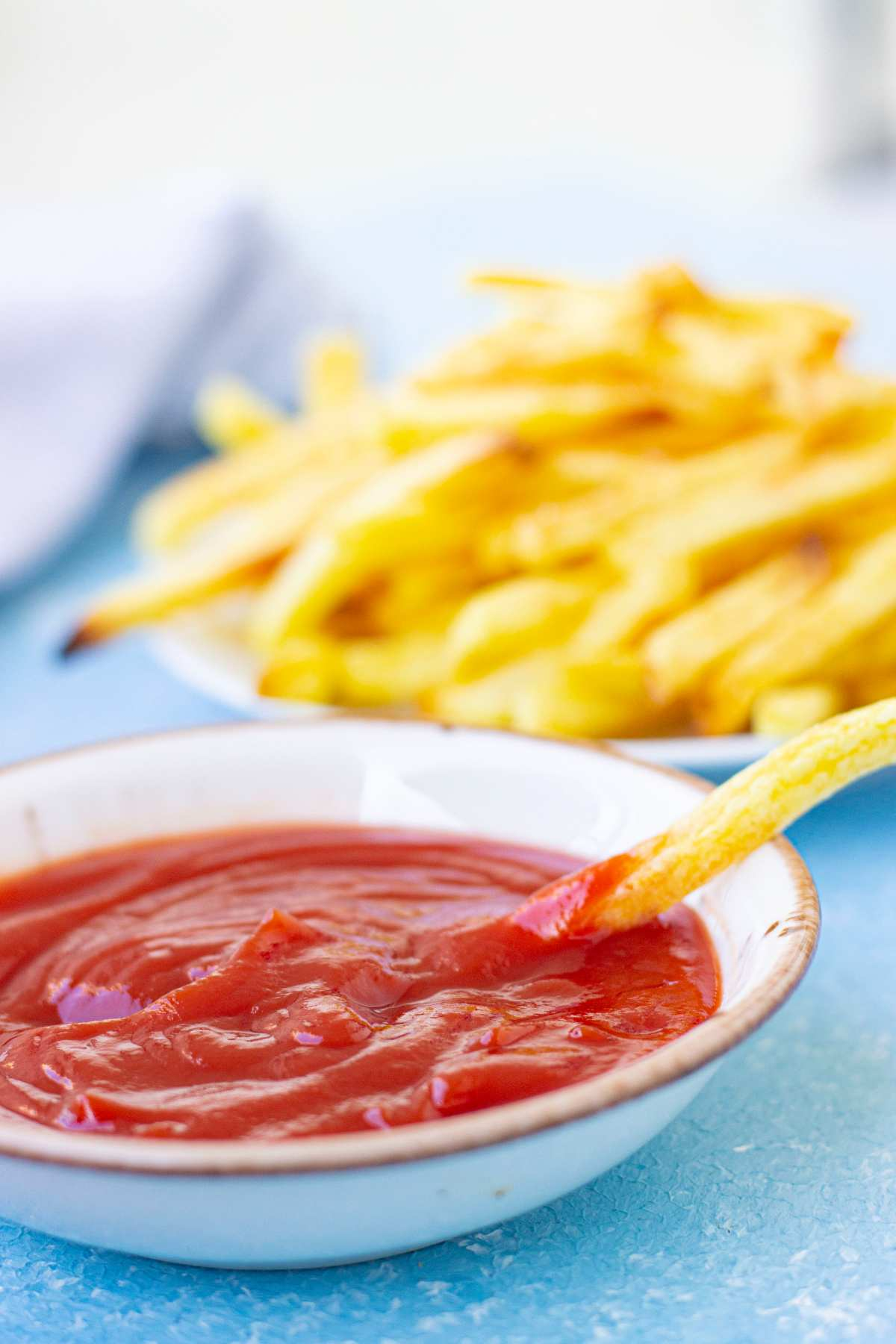 A bowl of ketchup with a fries being dipped with another bowl of cooked french fries at the back.