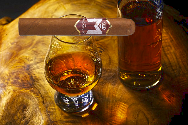 A bottle of M Bourbon by Macanudo with a glass of bourbon and a cherry, for a sweet and flavorful infused cigar experience