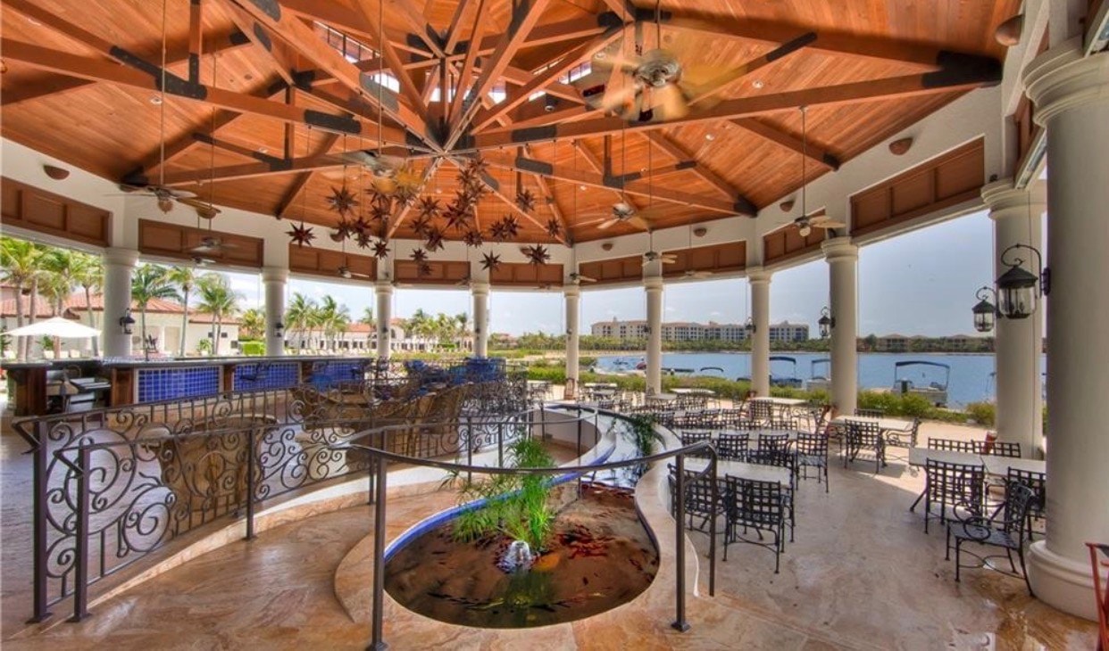 The image depicts a modern fitness center featuring large windows that provide a stunning view of the lake and the championship golf course at Miromar Lakes Beach and Golf Club. This luxurious community in Southwest Florida offers residents a vibrant lifestyle with access to amenities like a private white sand beach and gourmet dining options.