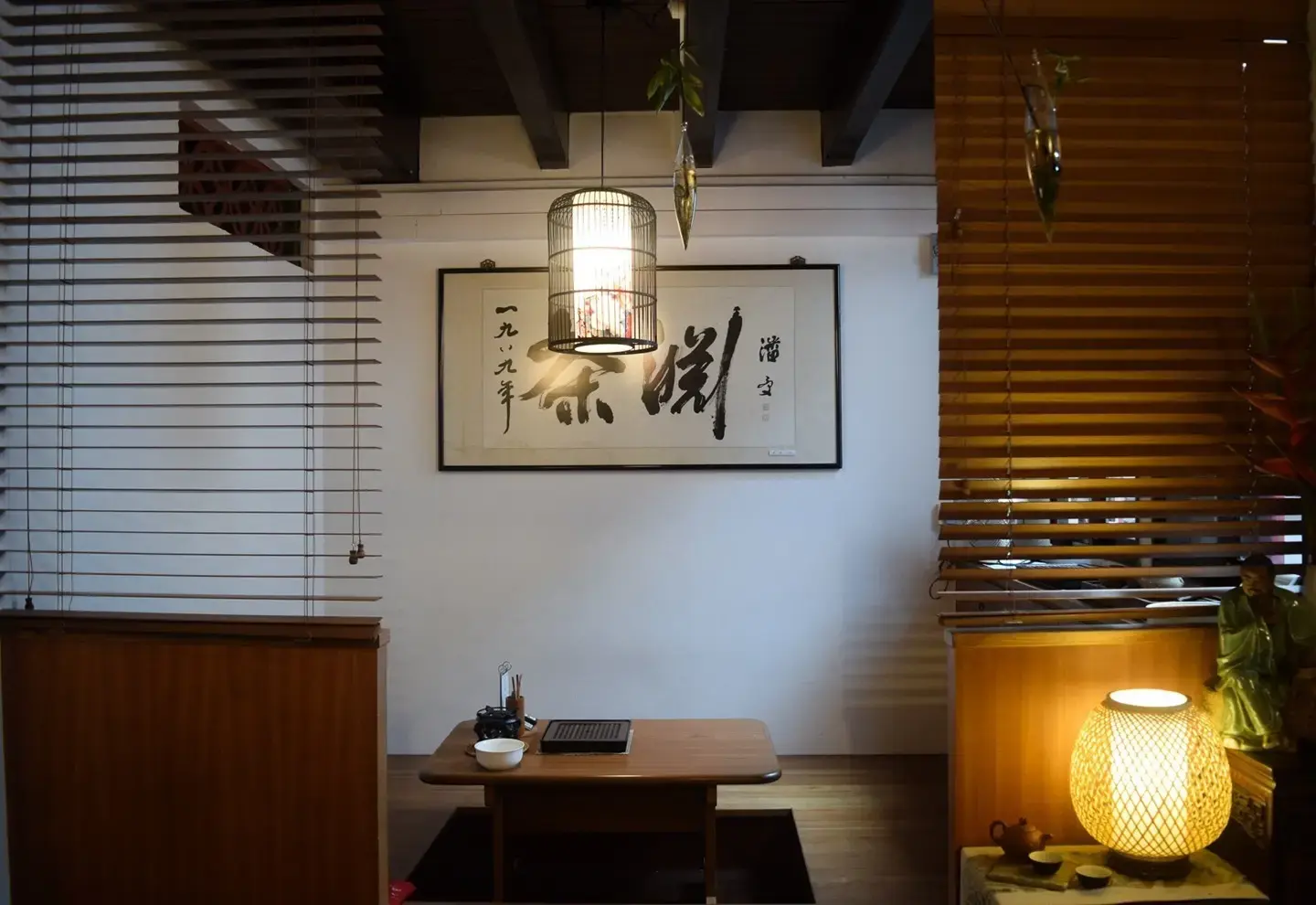 This image features a serene tea room with dark wooden furniture, warm lighting, and traditional East Asian decor. A calligraphy scroll hangs on the white wall behind a low table, framed by slatted wooden blinds on either side.