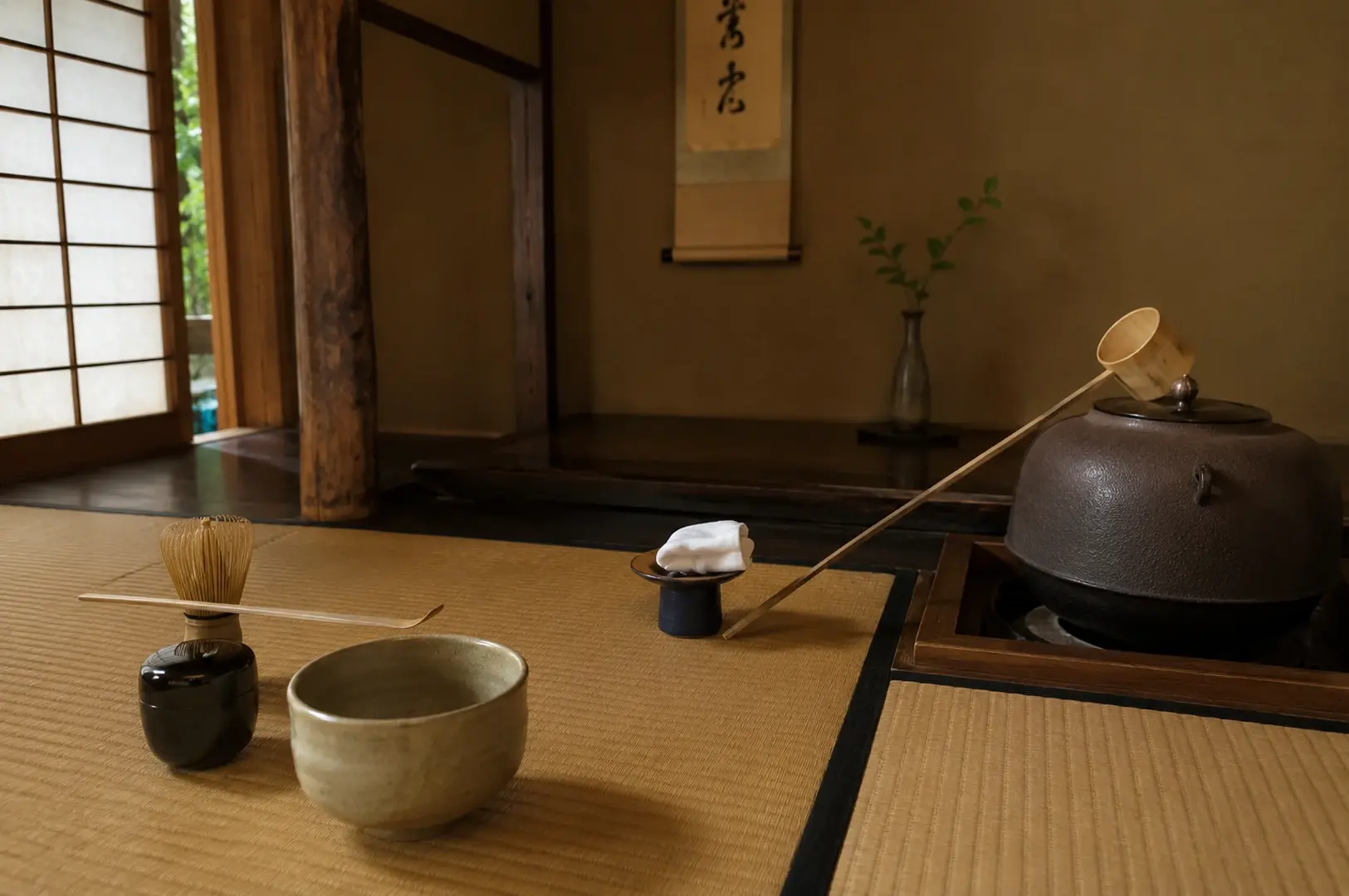 A quiet and minimalist view of a Japanese tea ceremony setup on tatami mats inside a traditional room. On the left, a light beige ceramic tea bowl, a small black lacquer caddy, and a bamboo whisk with a long scoop resting across it are neatly placed. To the right, a heavy iron kettle sits inside a wooden sunken hearth frame, with a long bamboo ladle resting diagonally against it. The background features clean earth-toned walls, a hanging calligraphy scroll, a slender vase with a green leafy branch, and an open wooden shoji doorway leading outside.