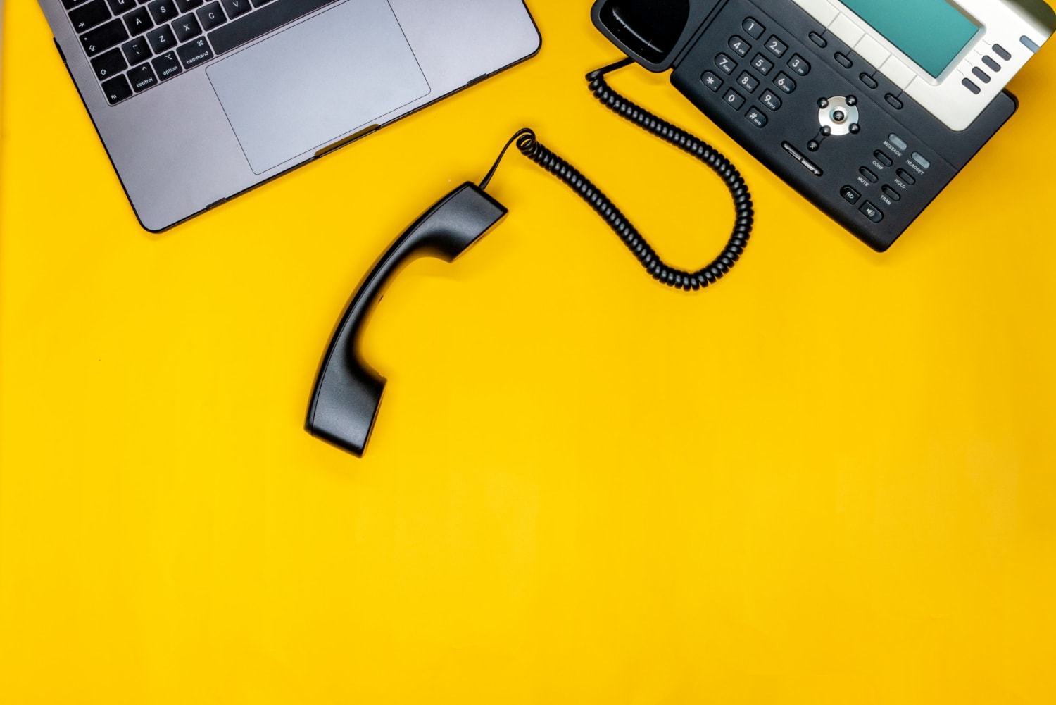 A business telephone with a detached handset on a vibrant yellow background, alongside a laptop. This setup conveys efficient Business VoIP providers supporting streamlined Cloud communications for remote and office professionals.