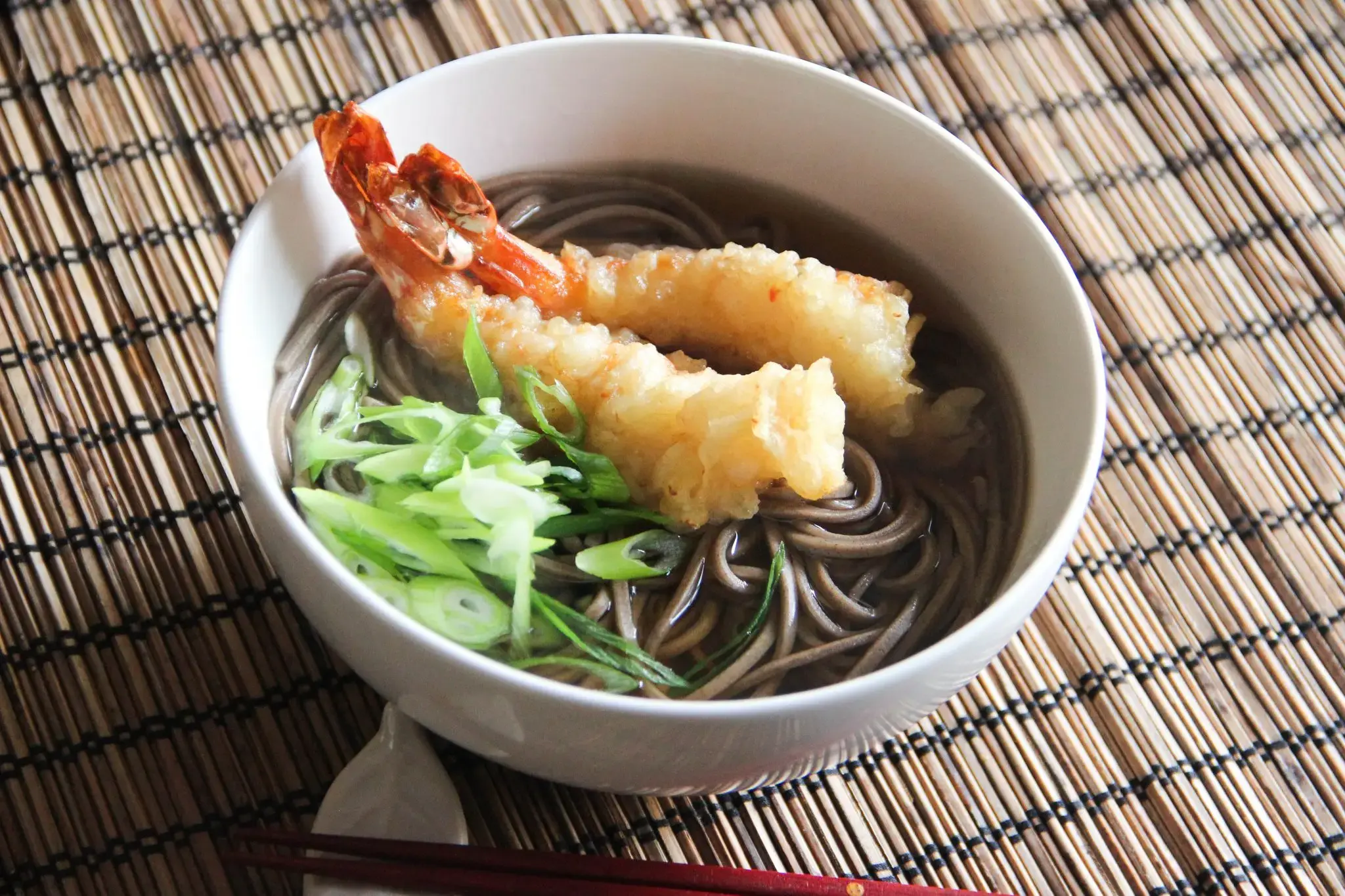 Two large, golden-fried shrimp tempura rest atop a bed of dark soba noodles submerged in a clear broth. The bowl is garnished with freshly sliced green onions and sits on a textured bamboo mat alongside a pair of dark red chopsticks.