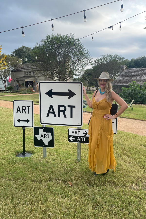Visitor in a yellow dress and hat standing beside directional art signs on a grassy lawn at Round Top, Texas, with historic buildings and string lights in the background.