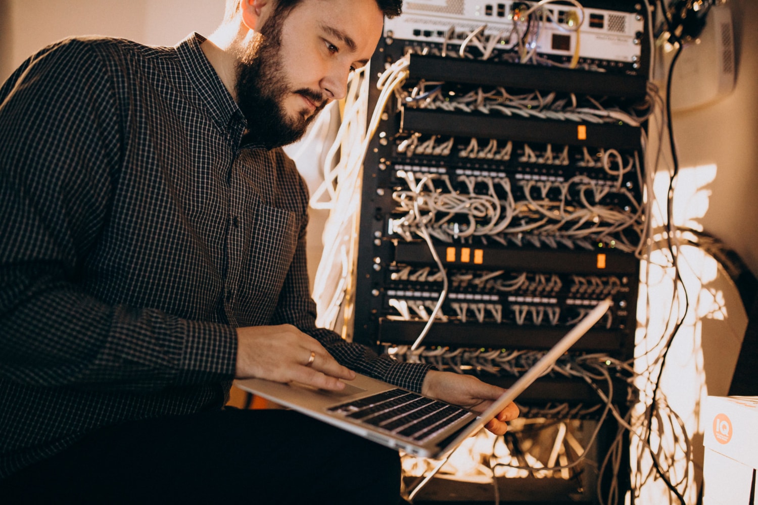 A bearded man working on a laptop in front of a server rack filled with tangled cables. He is setting up telecom security and managed telecom services for business phone systems and VoIP solutions.
