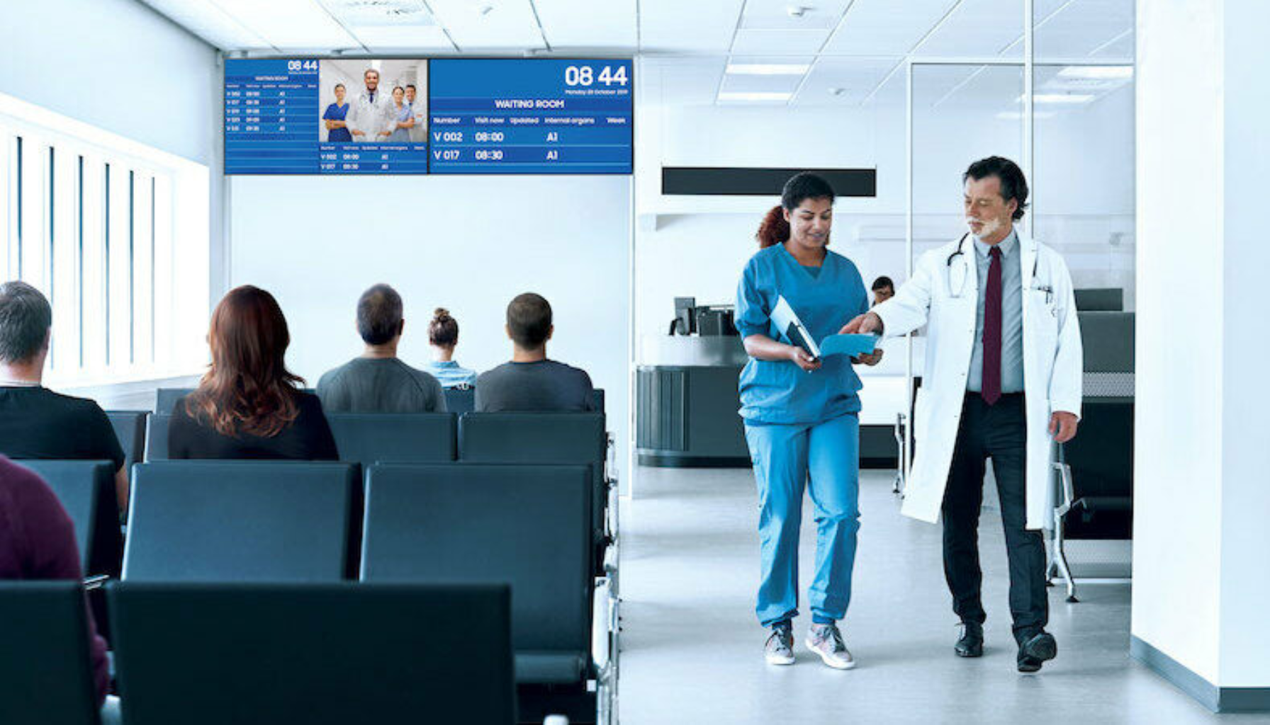 Pasienter venter i sykehusets venterom mens lege og sykepleier samarbeider foran skjermtavle.