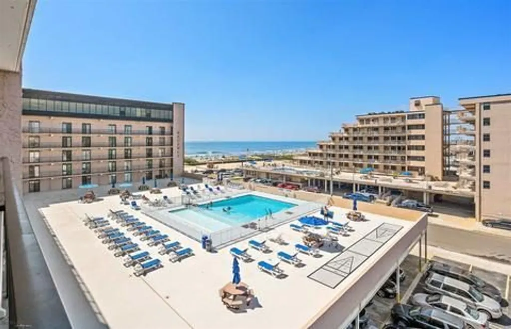 Elevated view of the Regency Towers pool deck in North Wildwood, NJ, featuring a large outdoor pool, lounge chairs, shuffleboard courts, and ocean views in the background.
