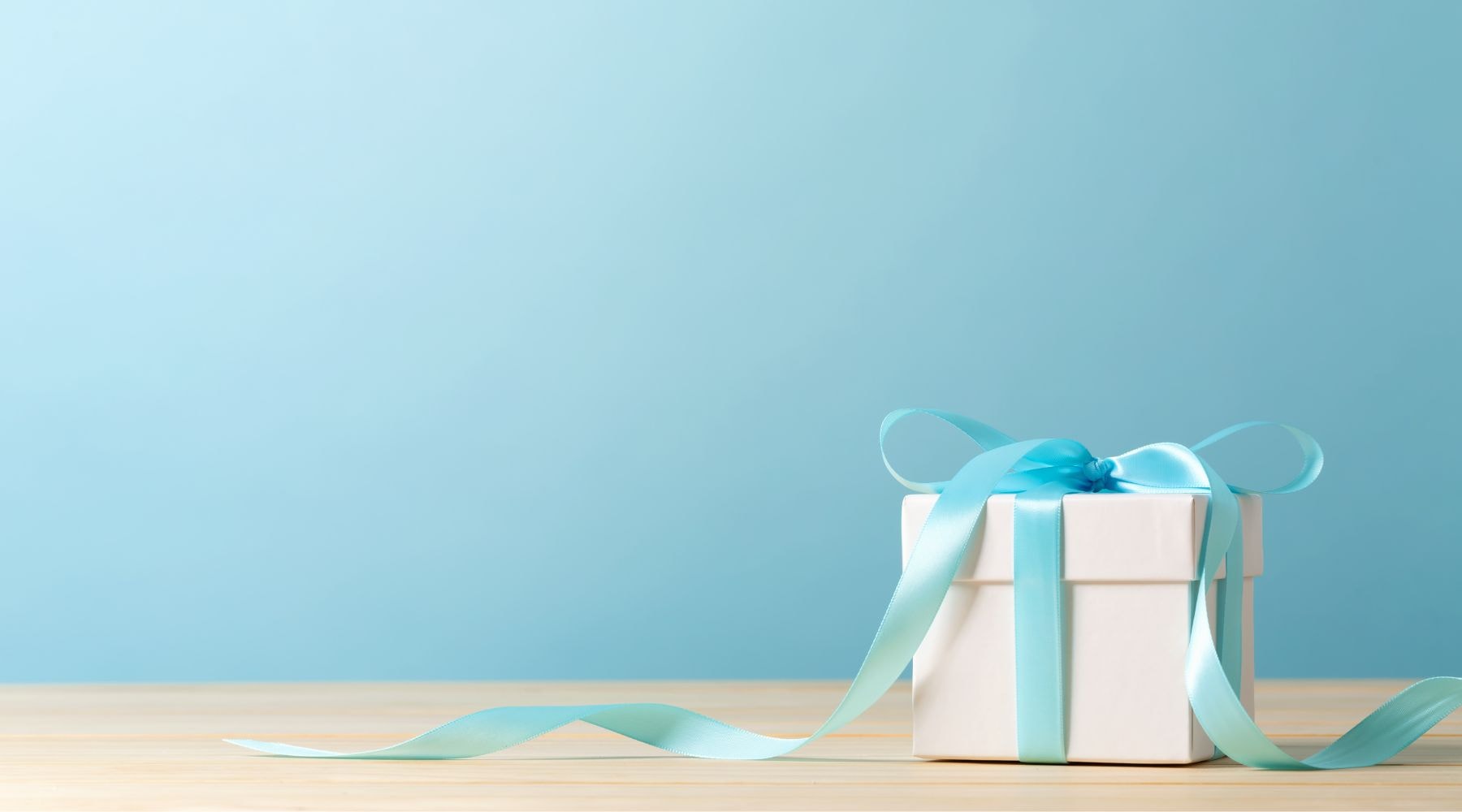 Simple white gift box with a teal ribbon on a wooden table against a soft blue background.